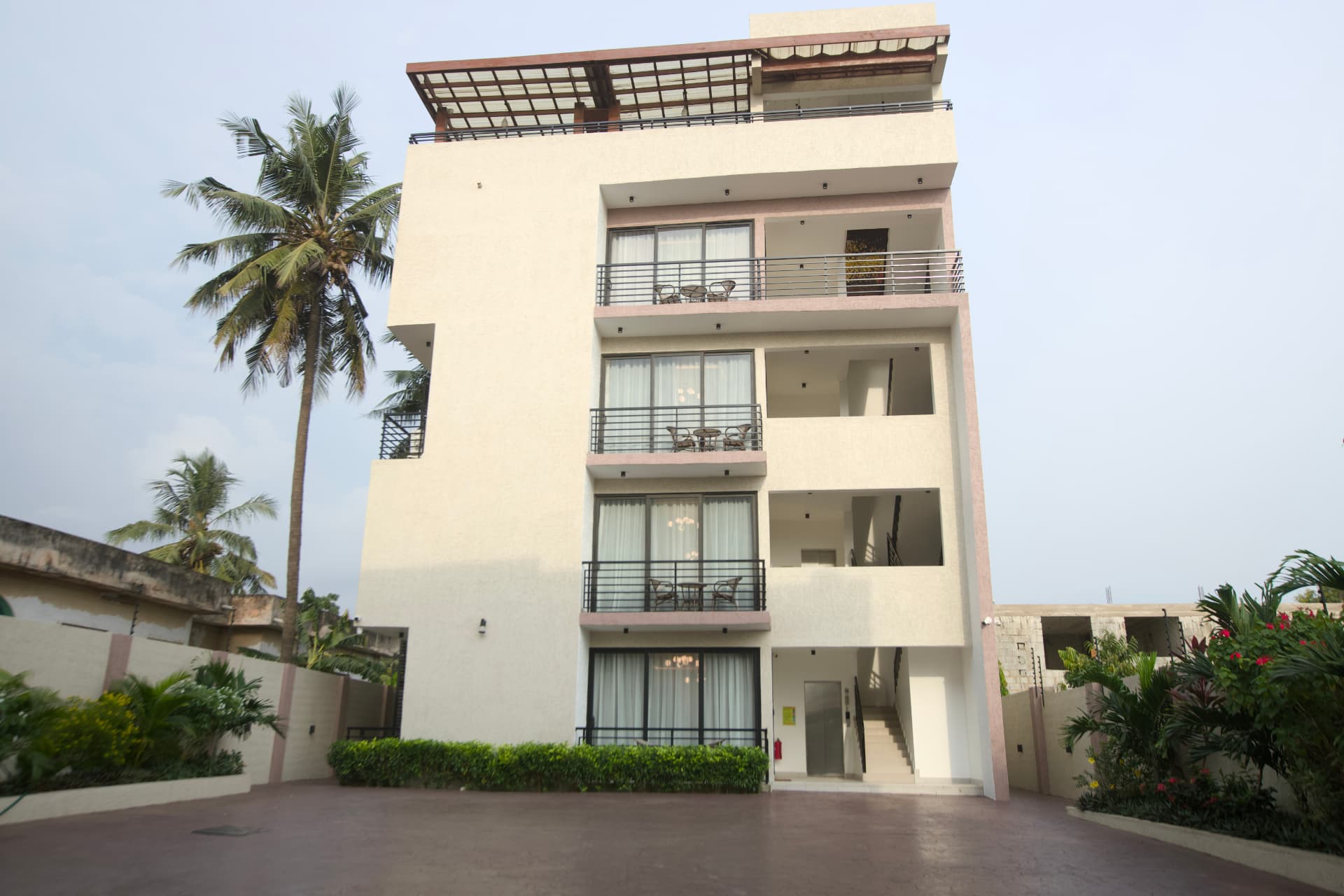 A cream multi-storey apartment building with furnished balconies, a rooftop pergola, and a palm tree at the entrance